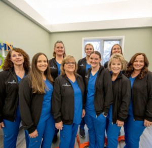 A group picture of the staff of Oral Surgery Associates of Lansing. They are wearing blue scrubs with striking black jackets branded with the practice's logo.